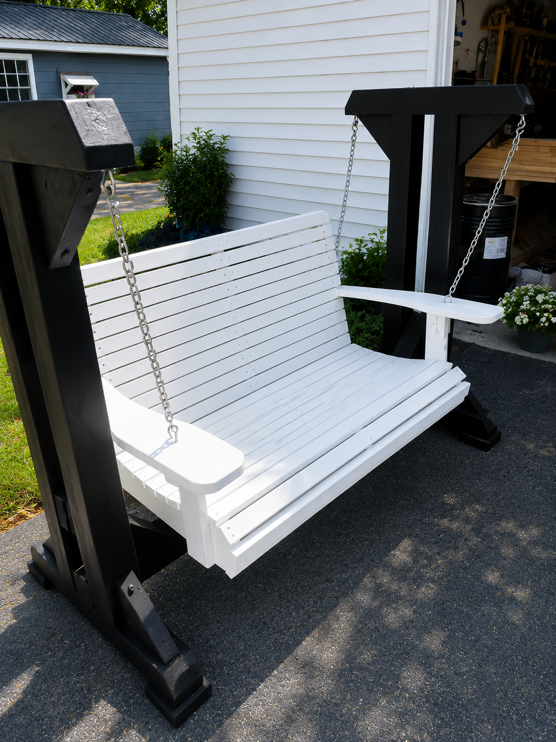 White-painted wooden porch swing with slatted seat and back, hanging by chains from a matching black-stained wood A-frame stand, positioned on a driveway beside a house.