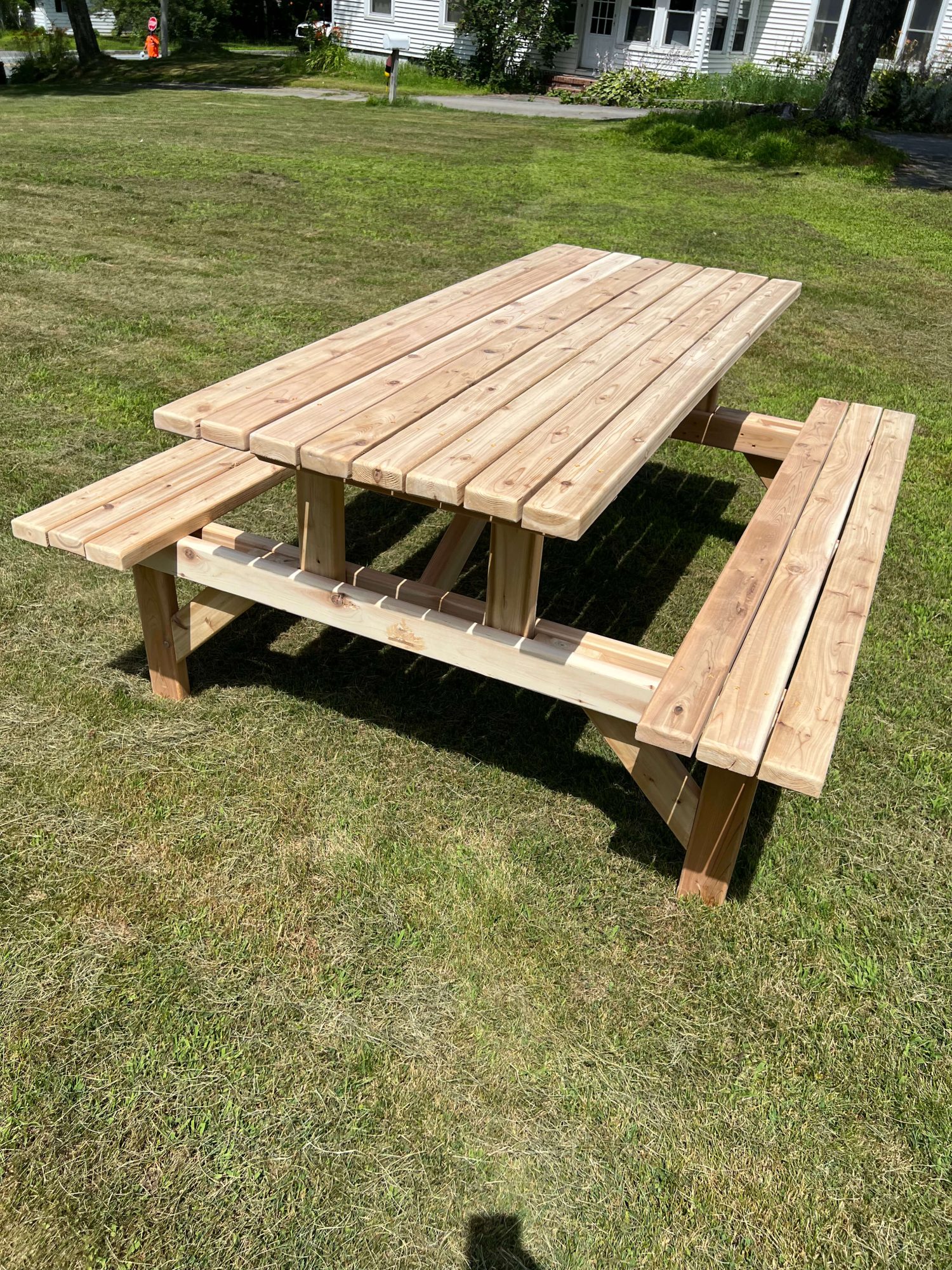 Rectangular cedar picnic table with two long attached benches, freshly built with visible wood grain, set on a grassy lawn near a house.
