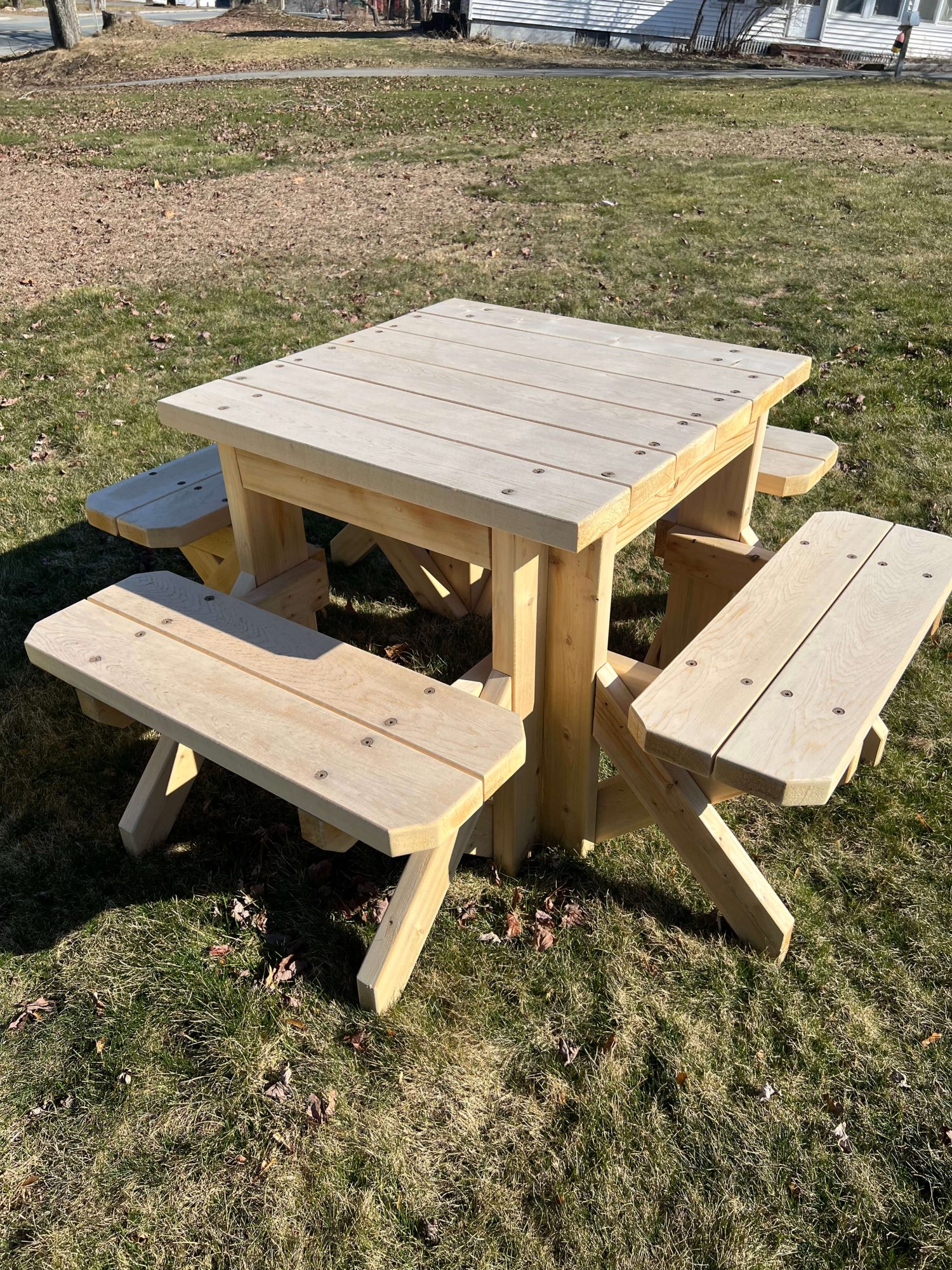 Handmade square wooden picnic table with four attached benches, set on a lawn. Unfinished light wood shows natural grain and joinery details.