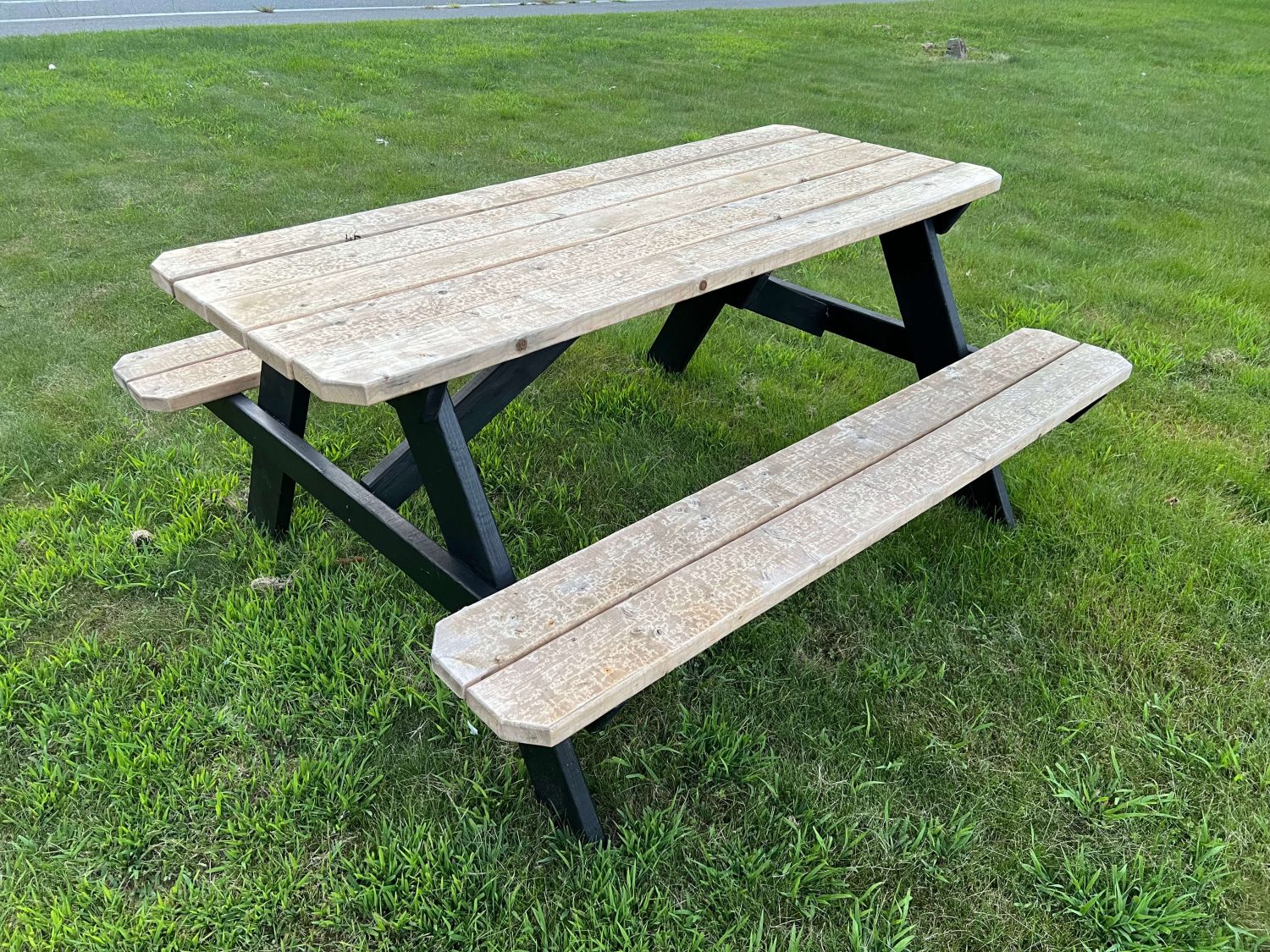 Classic A-frame picnic table with a black painted base and weathered natural wood top and benches, situated on grass near a road.