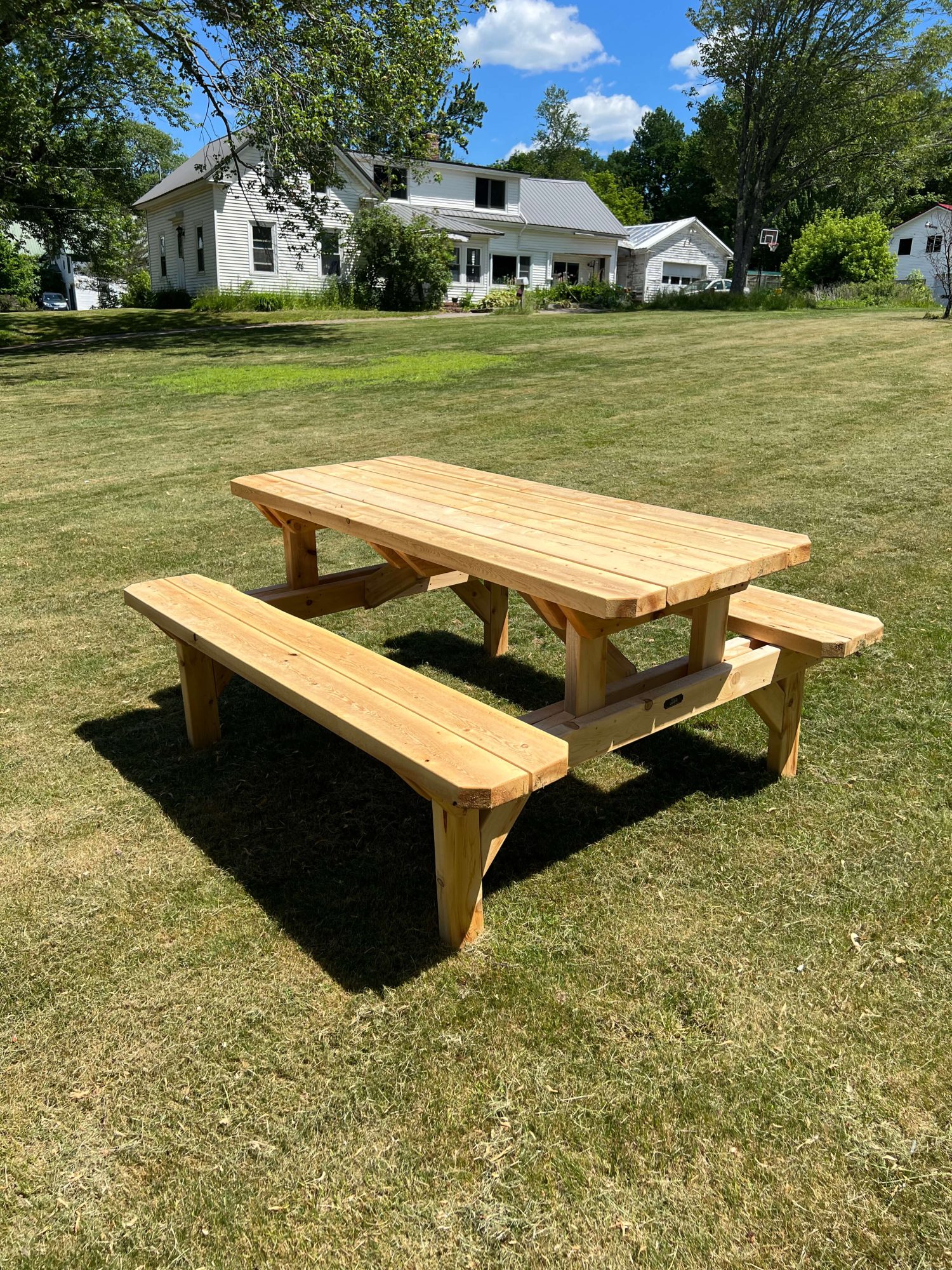 Rectangular wooden picnic table with two attached benches, natural finish, displayed on a lawn in front of a farmhouse with a basketball hoop in the background.