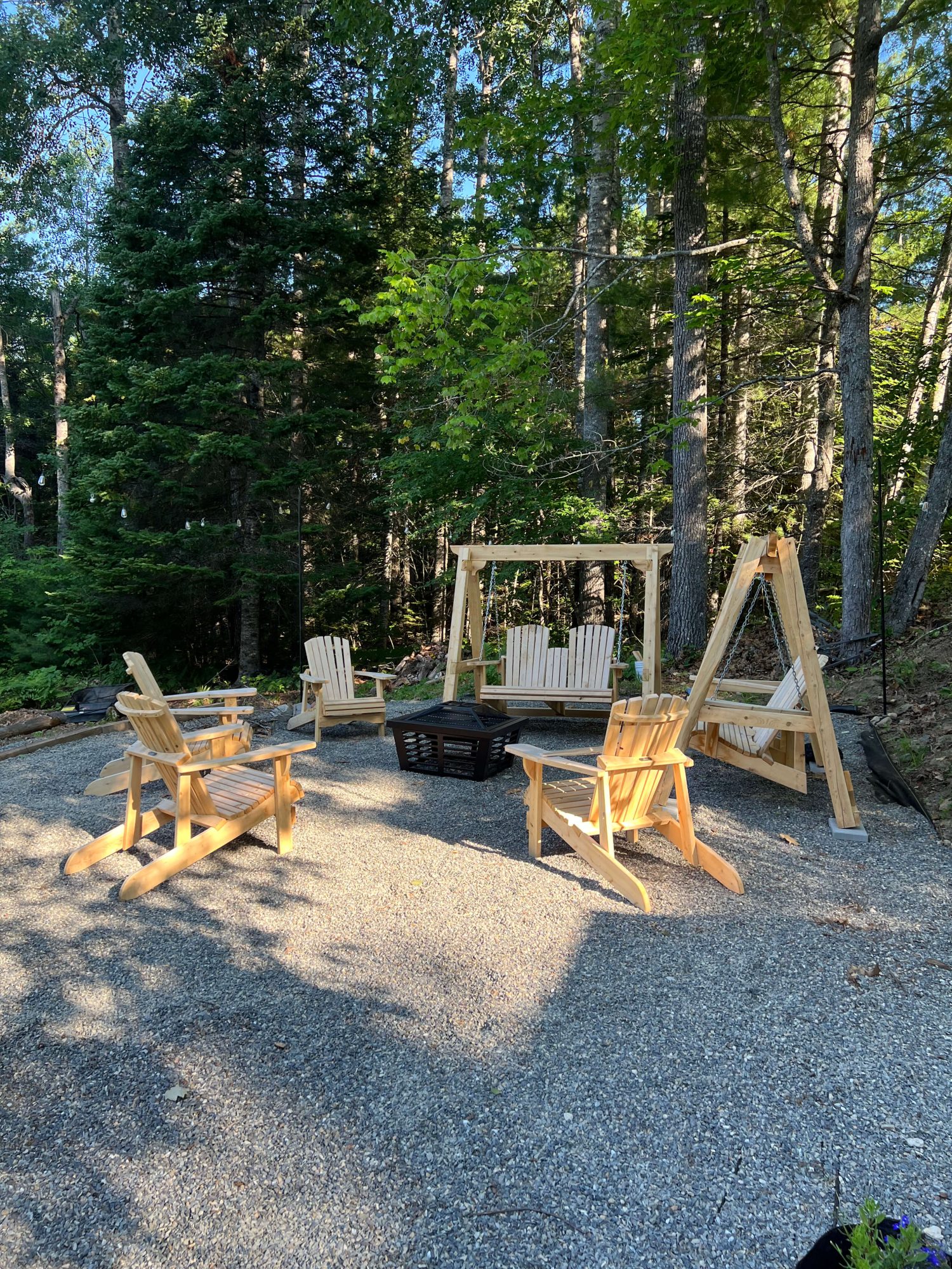 Outdoor seating area with a fire pit surrounded by four handmade Adirondack chairs and two A-frame porch swings with double Adirondack-style seats, set on a gravel patio in the woods.