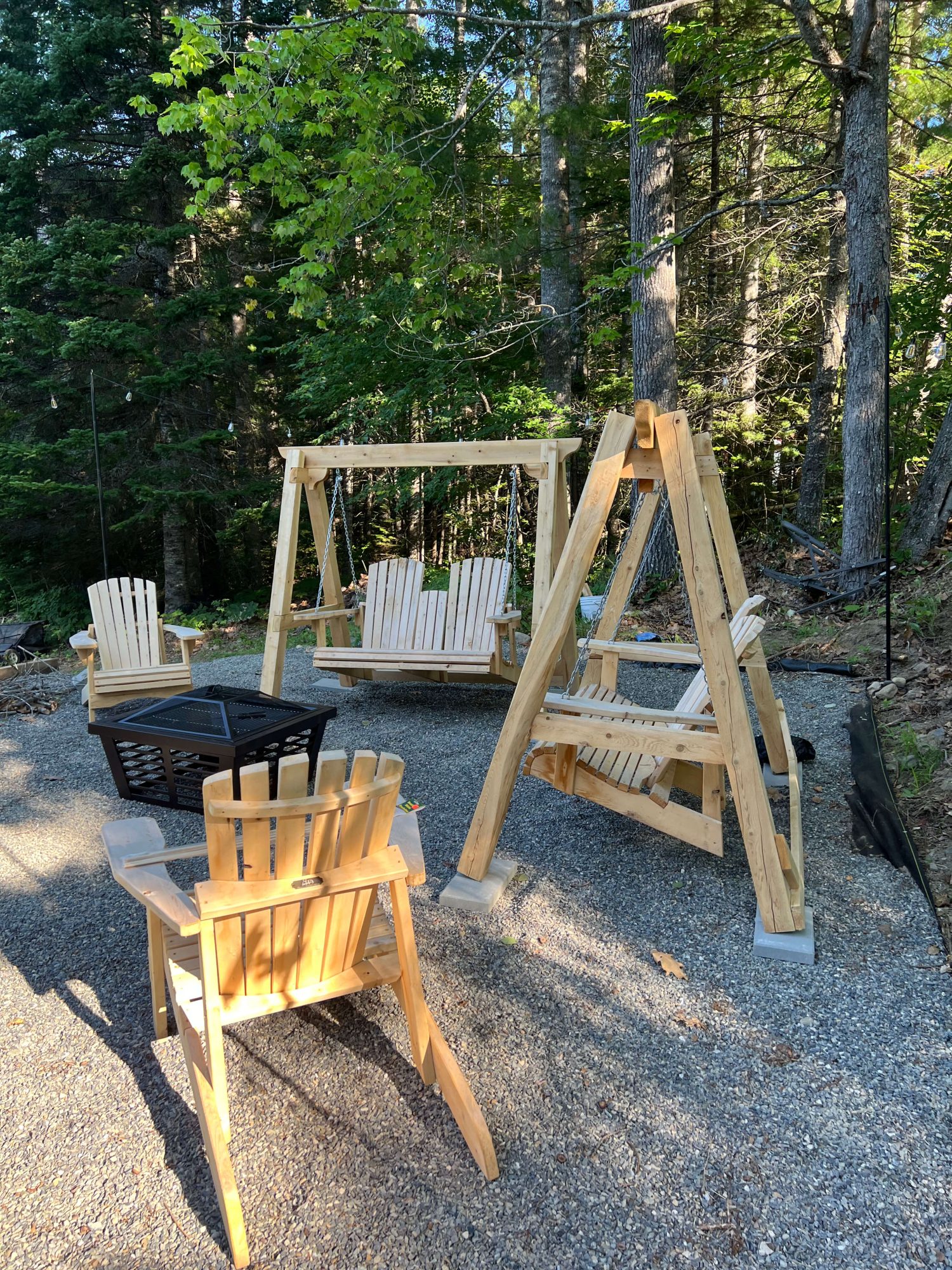 Two wooden A-frame porch swings with attached Adirondack-style bench seats, a black fire pit, and an Adirondack chair arranged on a gravel patio surrounded by trees.