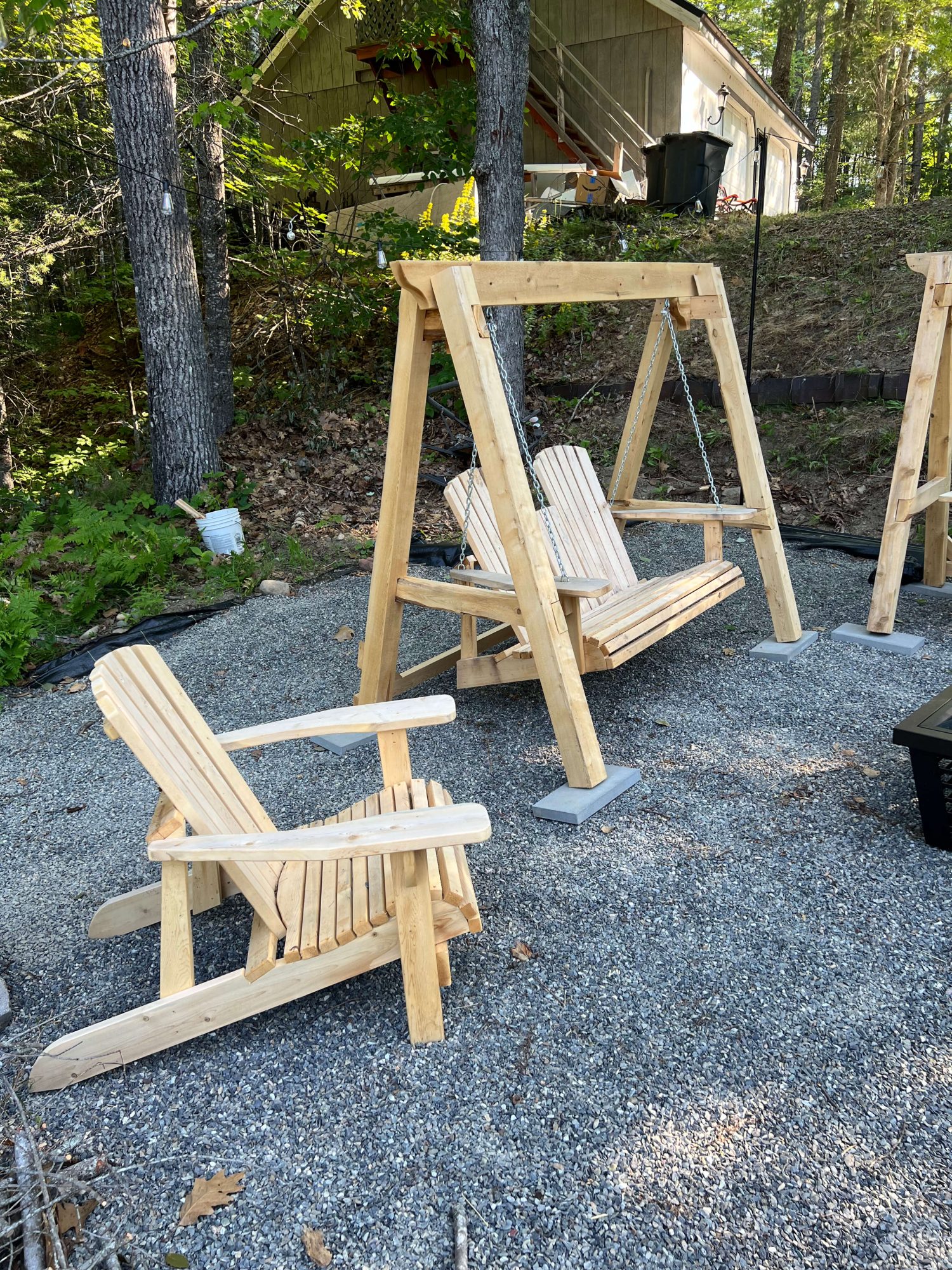 Wooden A-frame porch swing with a two-person Adirondack-style seat hanging by chains, alongside a matching single Adirondack chair on a gravel patio in a wooded setting.