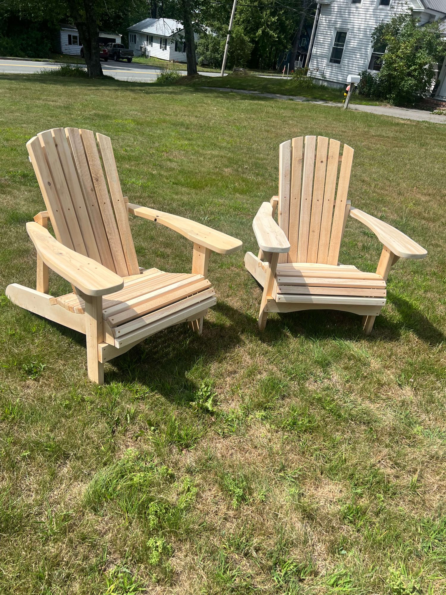 Pair of unfinished cedar Adirondack chairs with curved armrests and slatted backs, placed on a lawn near a residential street.