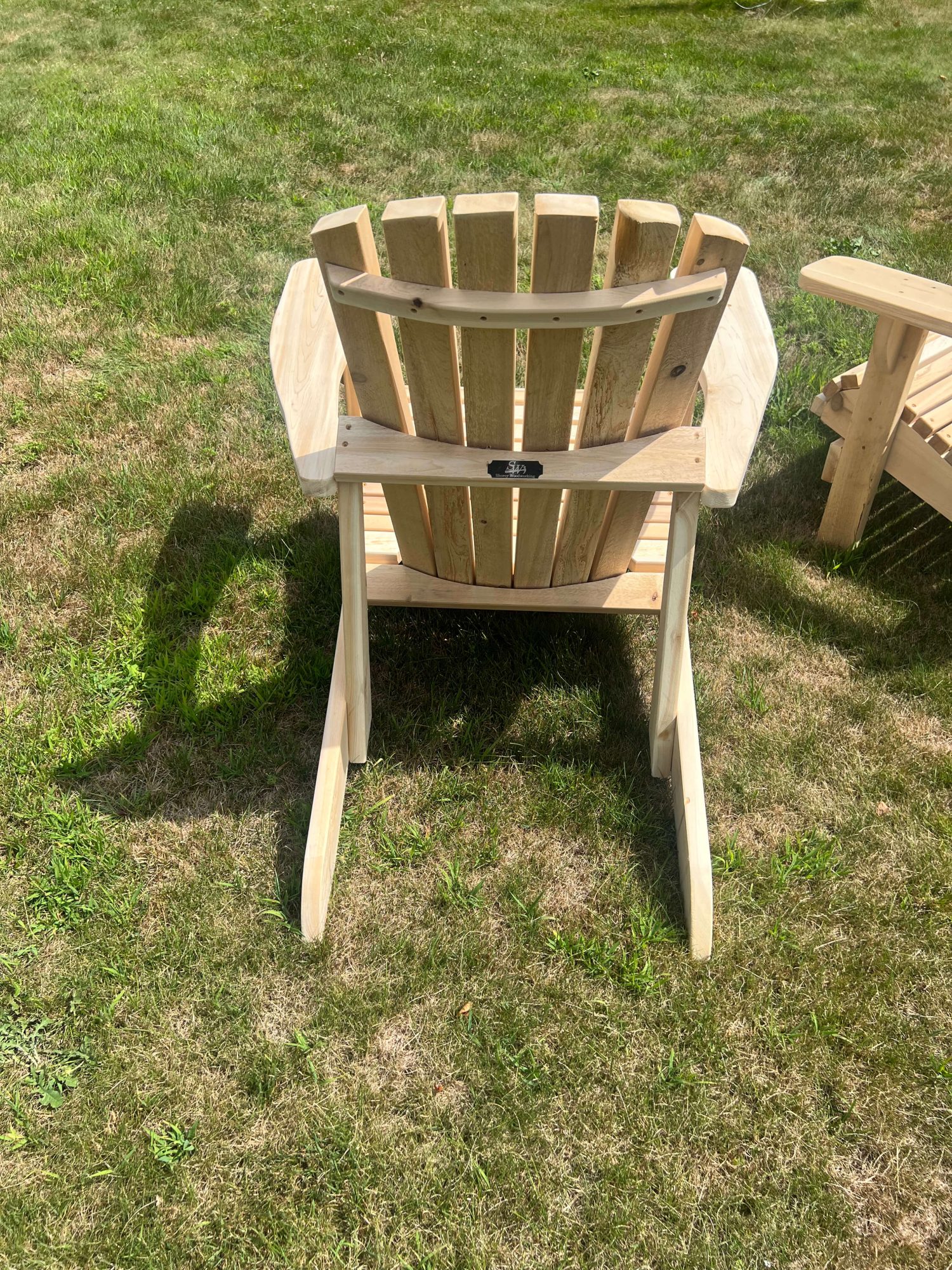 Handcrafted unfinished wood Adirondack chair shown from behind on a lawn, with a small Shorey Woodworking nameplate on the back crossbar; another chair visible in the background.